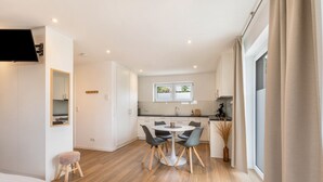 A modern hotel room with a bed, a small stool, a dining table with chairs, a kitchen area, and a large window.