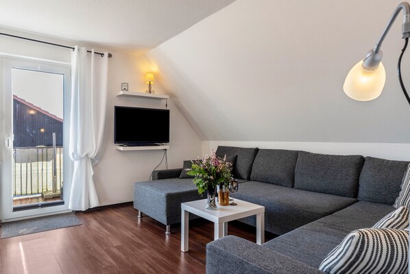 Un salon moderne avec un canapé gris, une table basse blanche, un téléviseur à écran plat fixé au mur et un balcon agrémenté d’un rideau blanc.