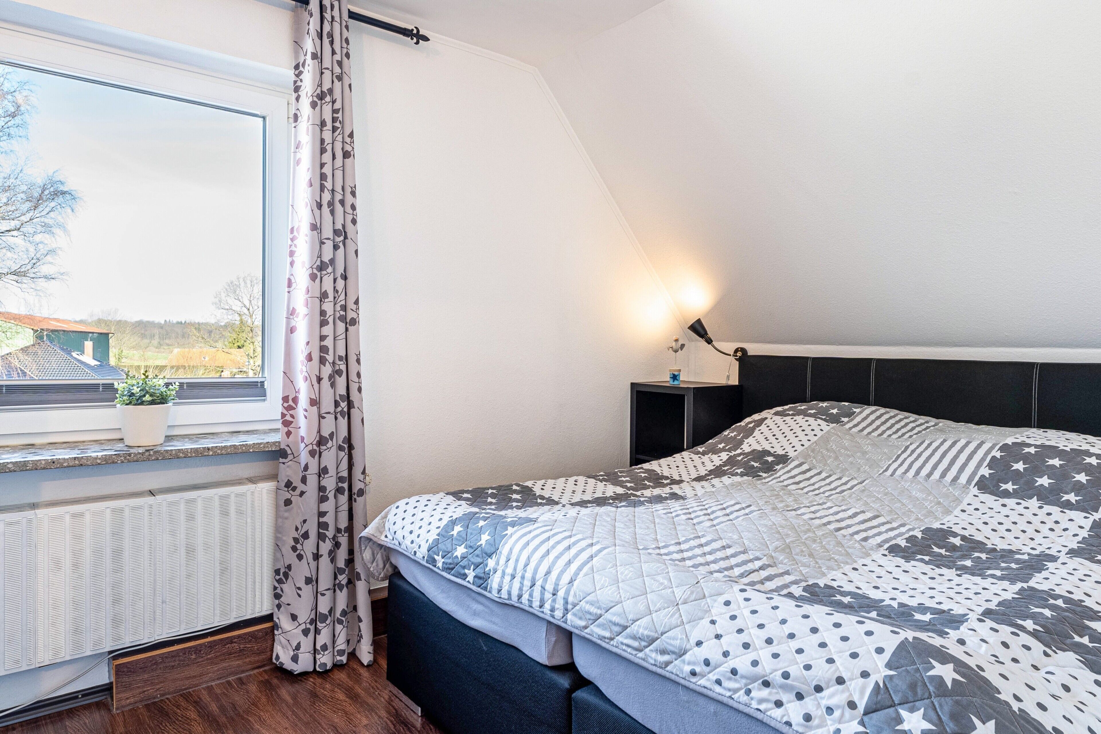 Un dormitorio con cama, ventana con cortinas, radiador, mesita de noche con lámpara y una planta en el alféizar.