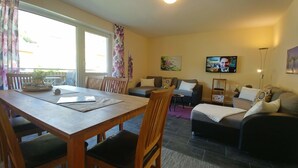 Ein Wohnzimmer mit einer Couch, einem Fernseher, einem Esstisch mit Stühlen und einem Fenster mit Vorhängen.