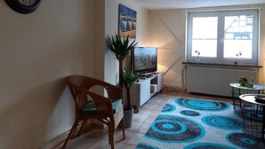 Una sala de estar con televisor, un reloj, una planta, una silla y una ventana con vista.