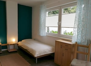 A small, single-bed room with a window, a bedside table, a wooden wardrobe, and a chair.