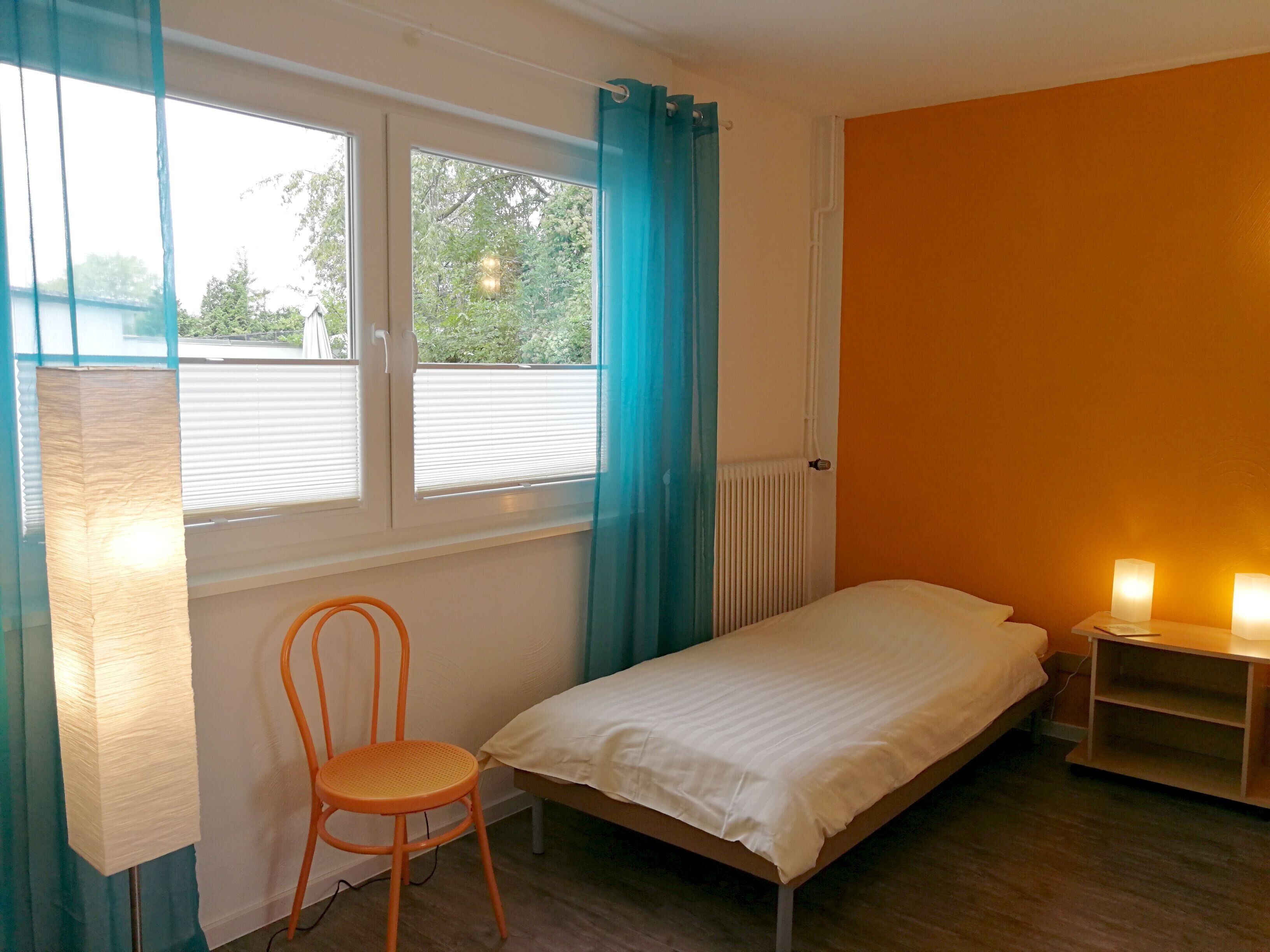 A bedroom with a single bed, a chair, a window with curtains, and a radiator.