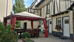 A covered outdoor seating area with a table and benches, surrounded by potted plants and a building with exposed timber framing. - Holiday Apartment 'Ferienhaus Rheinblick Wohnung 4' with Shared Terrace, Wi-Fi and Air Conditioning (Koblenz)