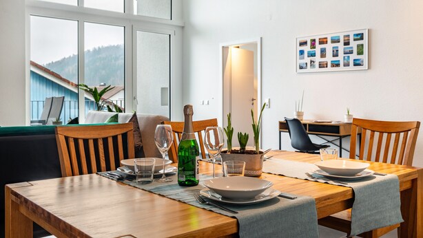 Ein Esszimmer mit Tisch, Geschirr, Gläsern und einer Flasche, umgeben von Stühlen, mit einem Fenster und einer gerahmten Fotocollage an der Wand.