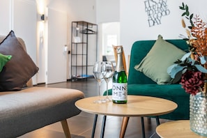 Un salon moderne avec un canapé vert, une table ronde en bois sur laquelle se trouve une bouteille de champagne et deux verres, ainsi qu'un vase décoratif rempli de fleurs.