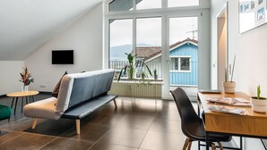 Ein modernes Wohnzimmer mit einer Couch, einem Esstisch, Stühlen und einem großen Fenster mit Blick auf ein blaues Haus und Berge.