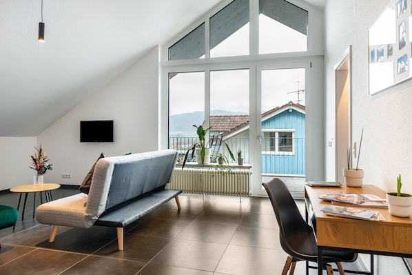Un salon moderne comprenant un canapé, une table à manger, des chaises et une grande fenêtre donnant sur une maison bleue et des montagnes.