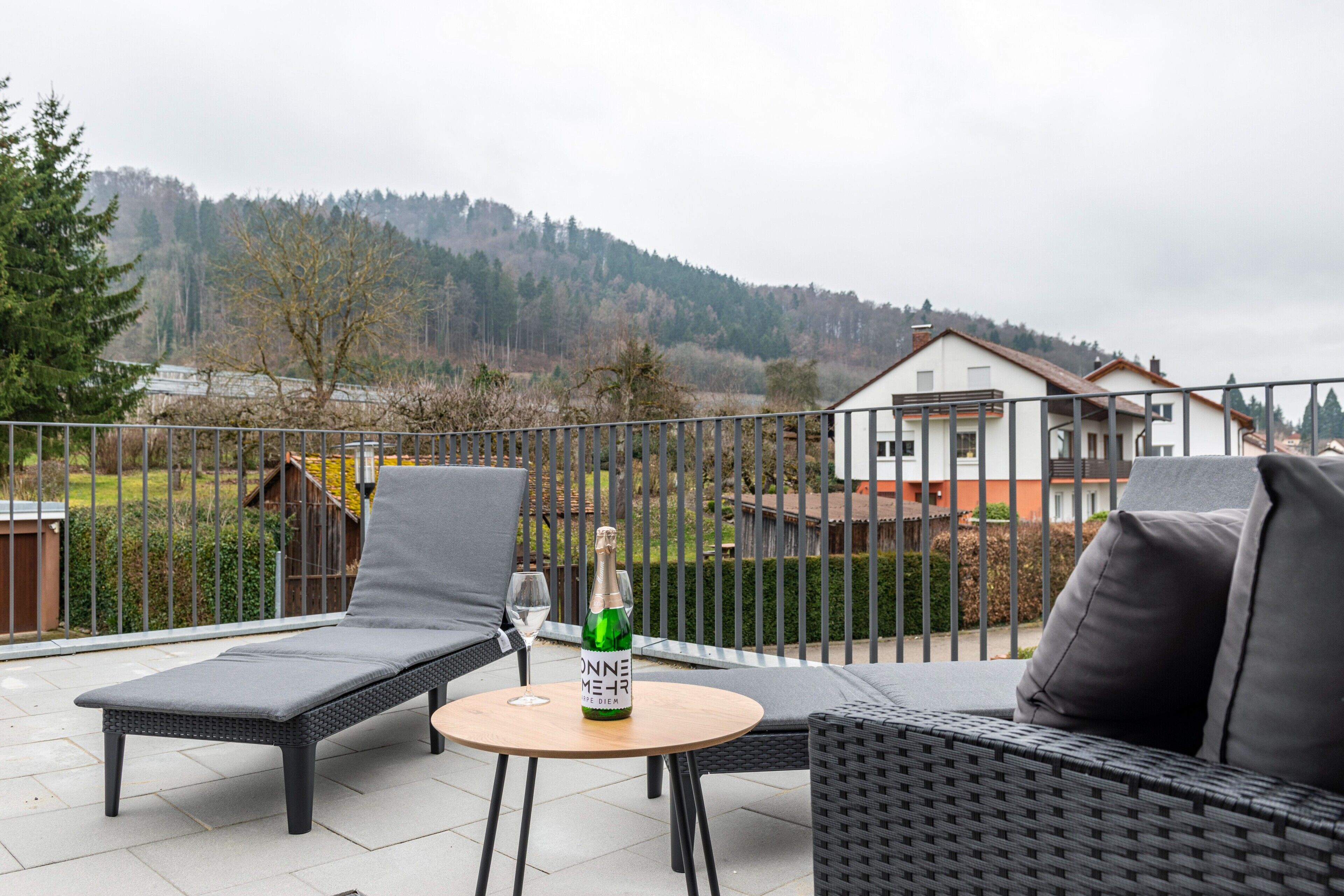 A balcony with outdoor furniture, a bottle of champagne, and a view of a house and forest.