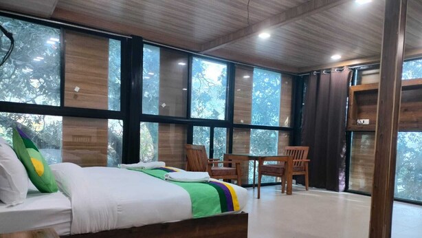 A modern bedroom with a large bed, wooden headboard, and a view of trees through large windows. - Hotel Lake In Tree (Nainital)