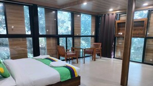 A modern bedroom with a wooden bed, a small table, two chairs, and large windows offering a view of trees. - Hotel Lake In Tree (Nainital)