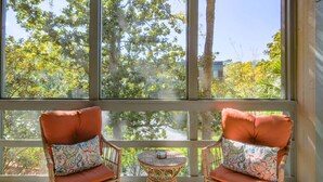 Deux fauteuils en osier avec des coussins orange et des oreillers à motifs, une petite table en osier et une grande fenêtre donnant sur des arbres et des bâtiments.