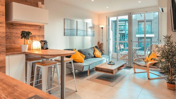 A modern living room with a sofa, coffee table, and chairs. There is a kitchen area with a counter and stools. - Modern 2BR 2BTH | Hypercenter | Croisette | AC (Cannes)
