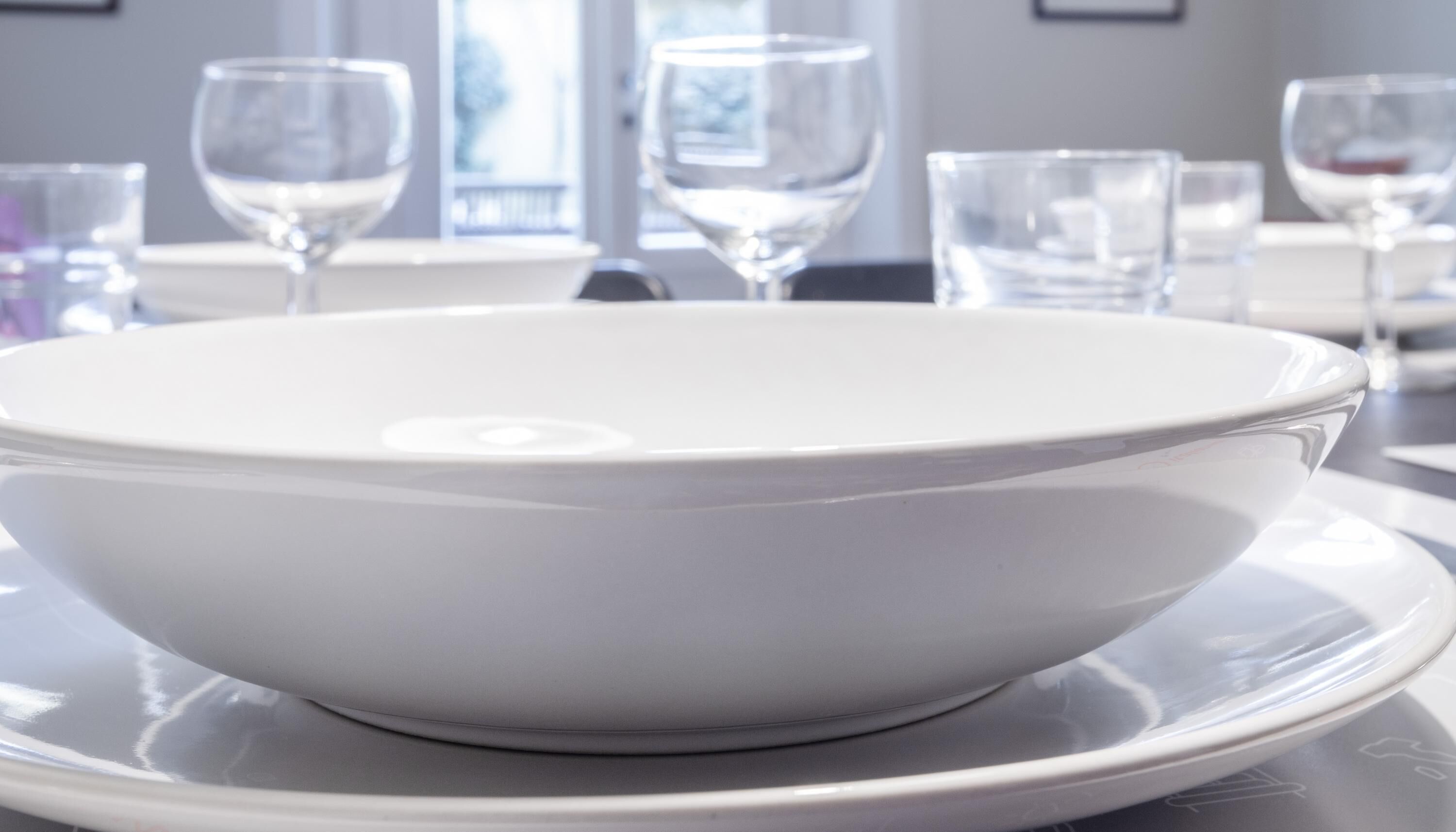 A white bowl on a white plate, with several wine glasses in the background.