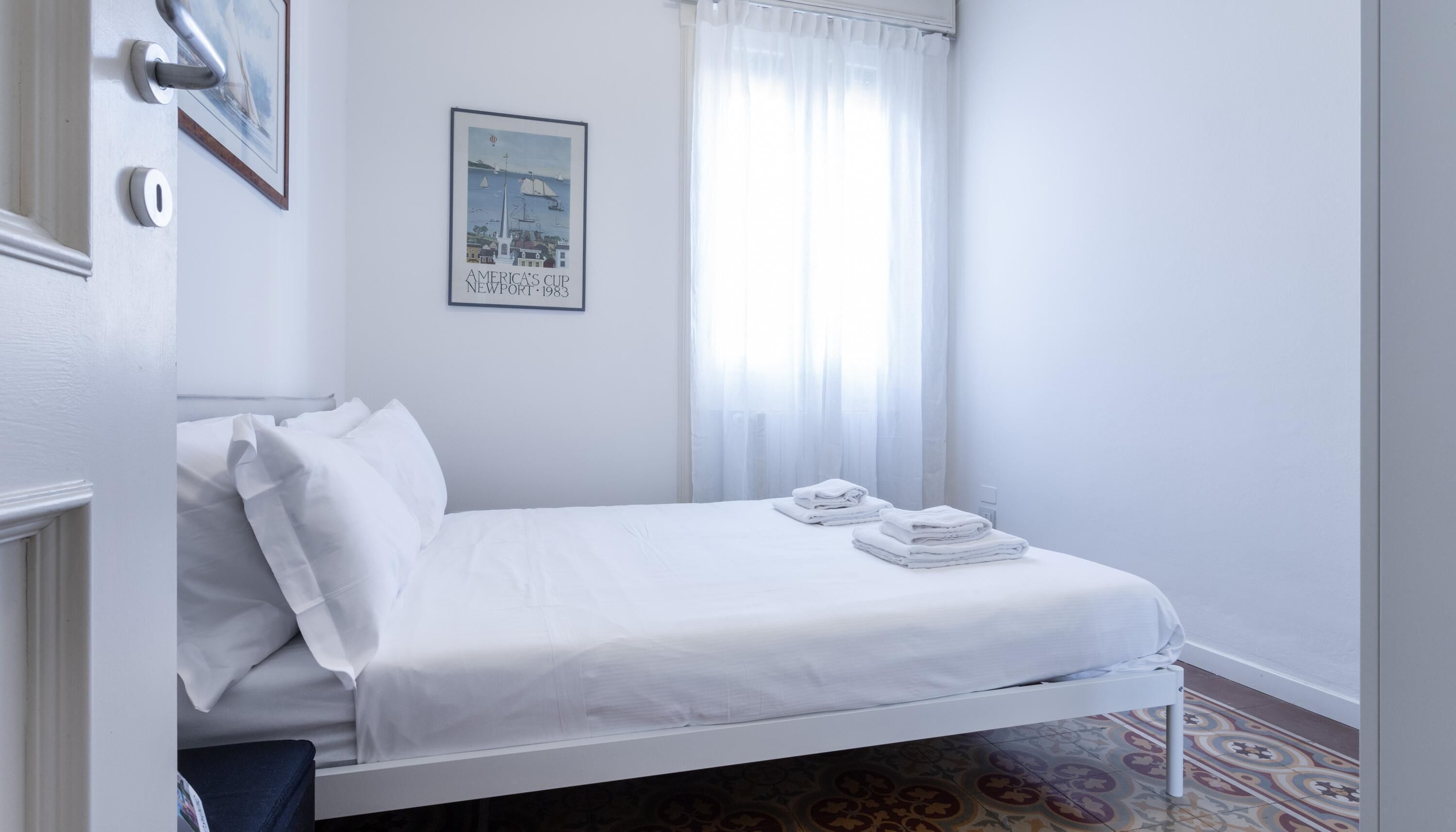 A neatly made bed with white linens and towels, a framed picture on the wall, and a window with sheer curtains.