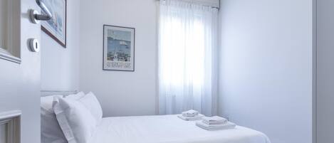 A neatly made bed with white linens and towels, a framed picture on the wall, and a window with sheer curtains.