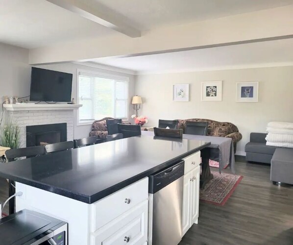 A modern kitchen with a central island, white cabinetry, and a built-in dishwasher. There is a fireplace, a flat-screen TV mounted above it, and a dining area with a table and chairs. The room has a large window, a sofa, and a rug.