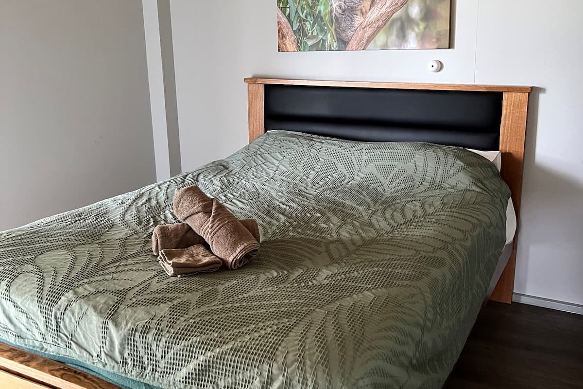 Un lit bien fait, avec une couvre-lit à motifs et une serviette enroulée, une tête de lit en bois et un tableau encadré représentant un koala accroché au mur.