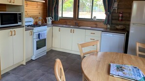 Pieni, käytännöllinen keittiö, jossa on valkoiset kaapit, mikroaaltouuni, liesi ja pesuallas. Keittiössä on ruokapöytä, jonka päällä on lehti, sekä ikkuna, jossa on verhot.