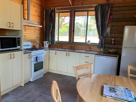 A compact kitchen with white cabinets, a microwave, stove, and sink. There's a dining table with a magazine, and a window with curtains.