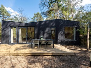 Une cabane moderne en bois sombre, dotée de grandes portes-fenêtres coulissantes, d’une terrasse en bois et d’une table avec des chaises.