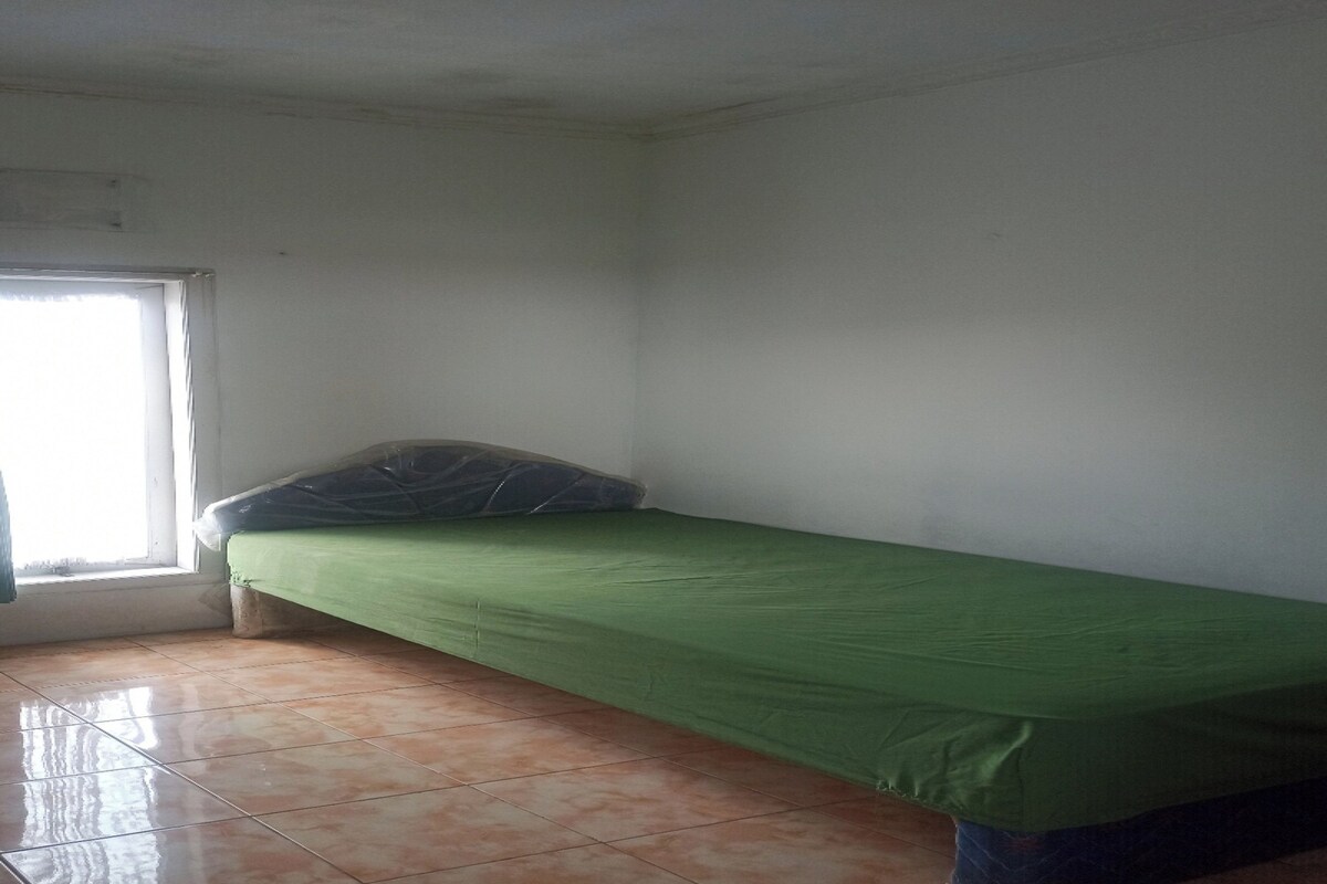 Ein kleines Zimmer mit einem Einzelbett, einer grünen Matratze, einem Fenster und Fliesenboden.