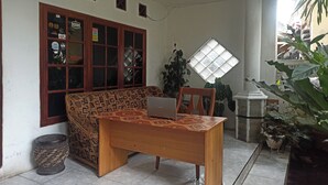 A room with a wooden desk, a patterned sofa, a window with a view, and a potted plant. - OYO Life 91339 Pondok Kost Aulia Syariah (Garut)