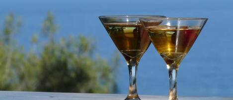 Two glasses of amber-colored beverage on a wooden surface with a blurred background of greenery and a body of water.