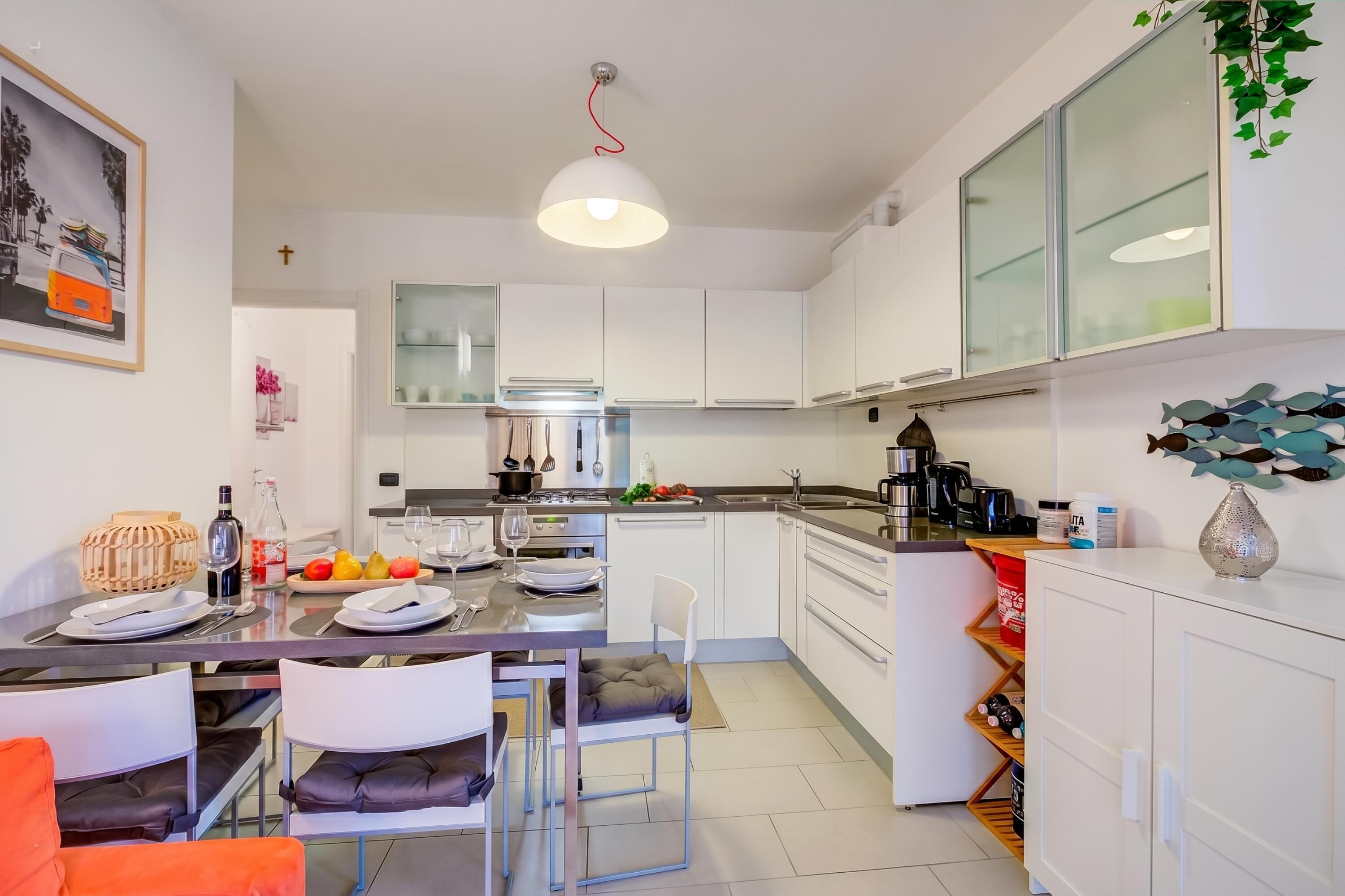 A modern kitchen with white cabinets, a dining area with chairs, a hanging light fixture, and a wall-mounted fish decoration.