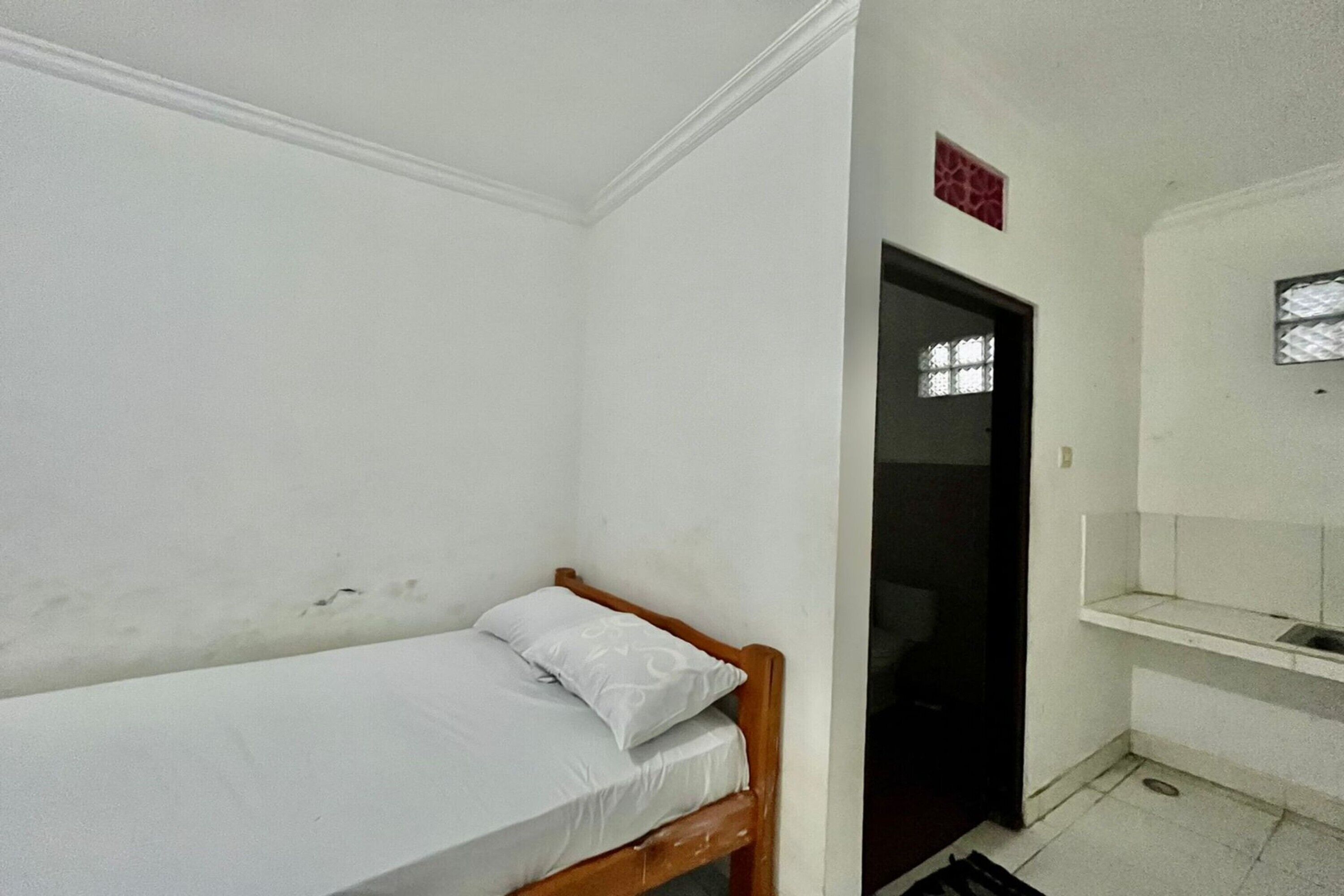 A small, single-bed room with a wooden frame and white bedding. A small shelf unit is visible to the right, and a door leads to another room.