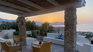 A covered outdoor seating area with wicker chairs and a white sofa, stone pillars, and a view of the sea at sunset. - Addauro Villa (Alcamo Marina)