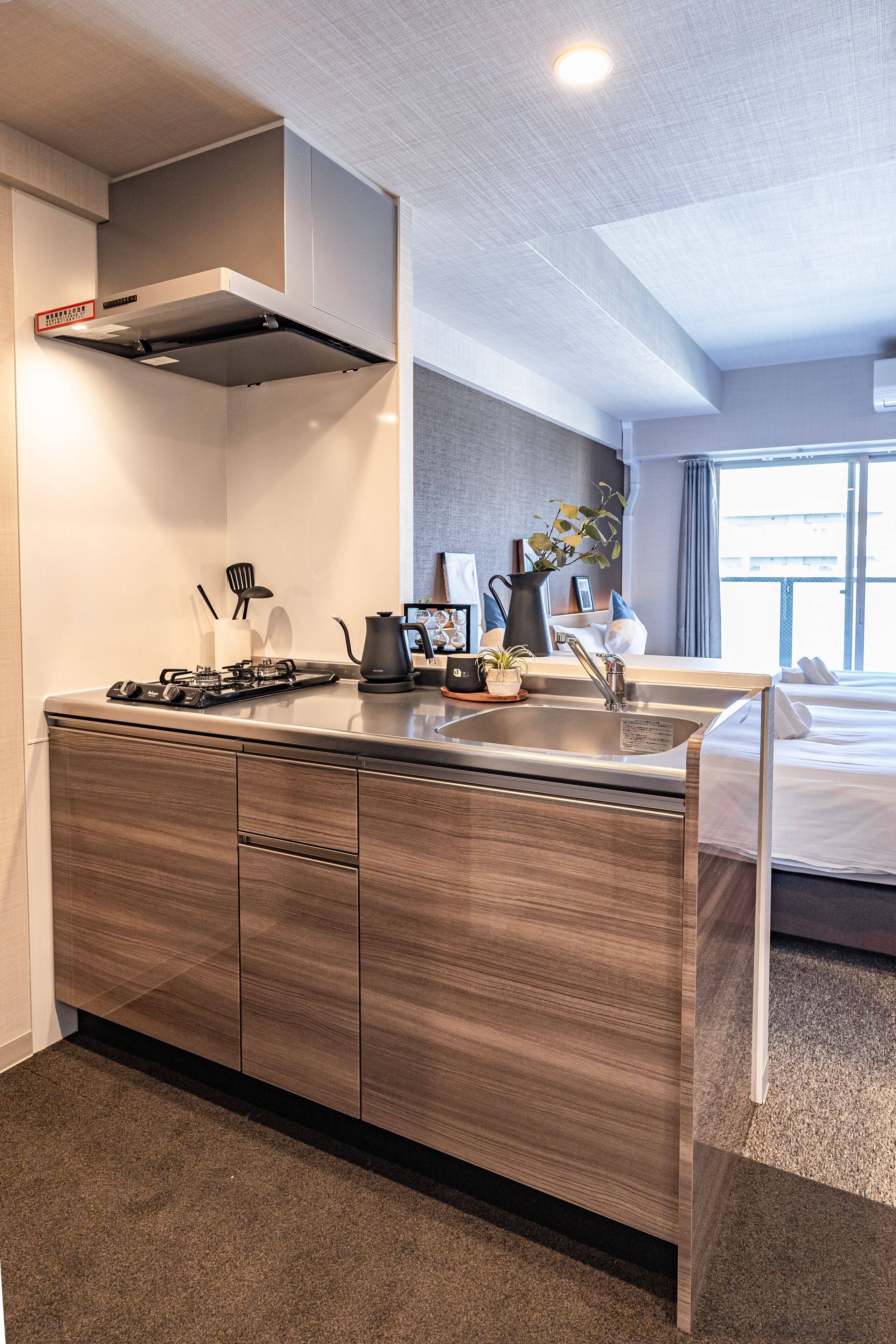 A modern hotel room with a kitchenette, a bed, a window with curtains, and a view of the city.