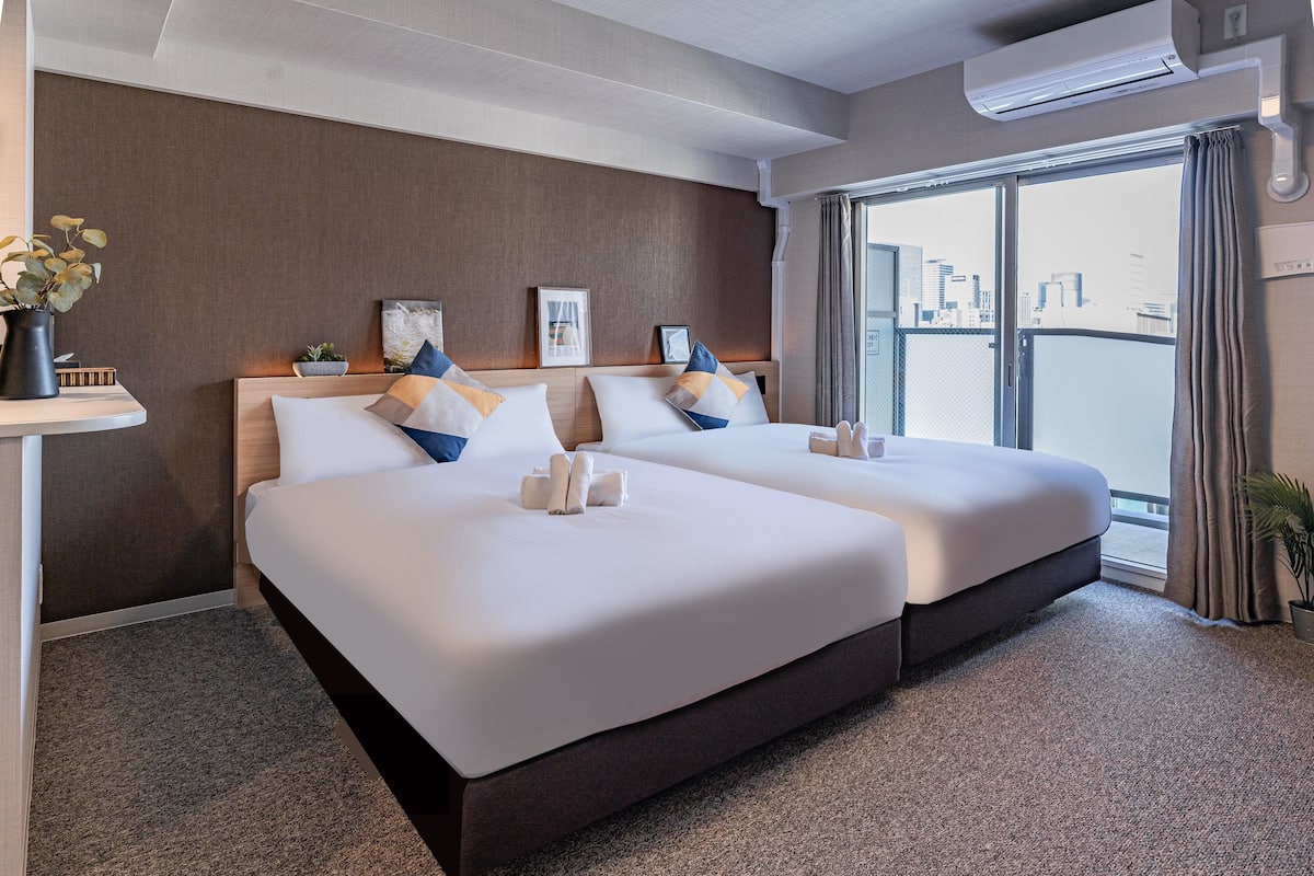 A hotel room with two beds, a city view, a plant, and a wall-mounted air conditioner.