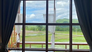 A room with a large window overlooking a green landscape with fields and a mountain in the distance. - Hinpha homestay (Pua)