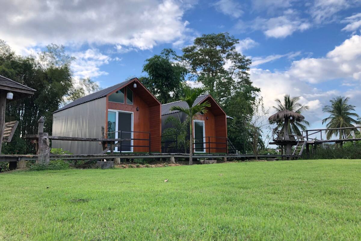 Un chalet rustique à l’extérieur métallique et aux éléments décoratifs en bois, entouré d’une végétation luxuriante et de palmiers.