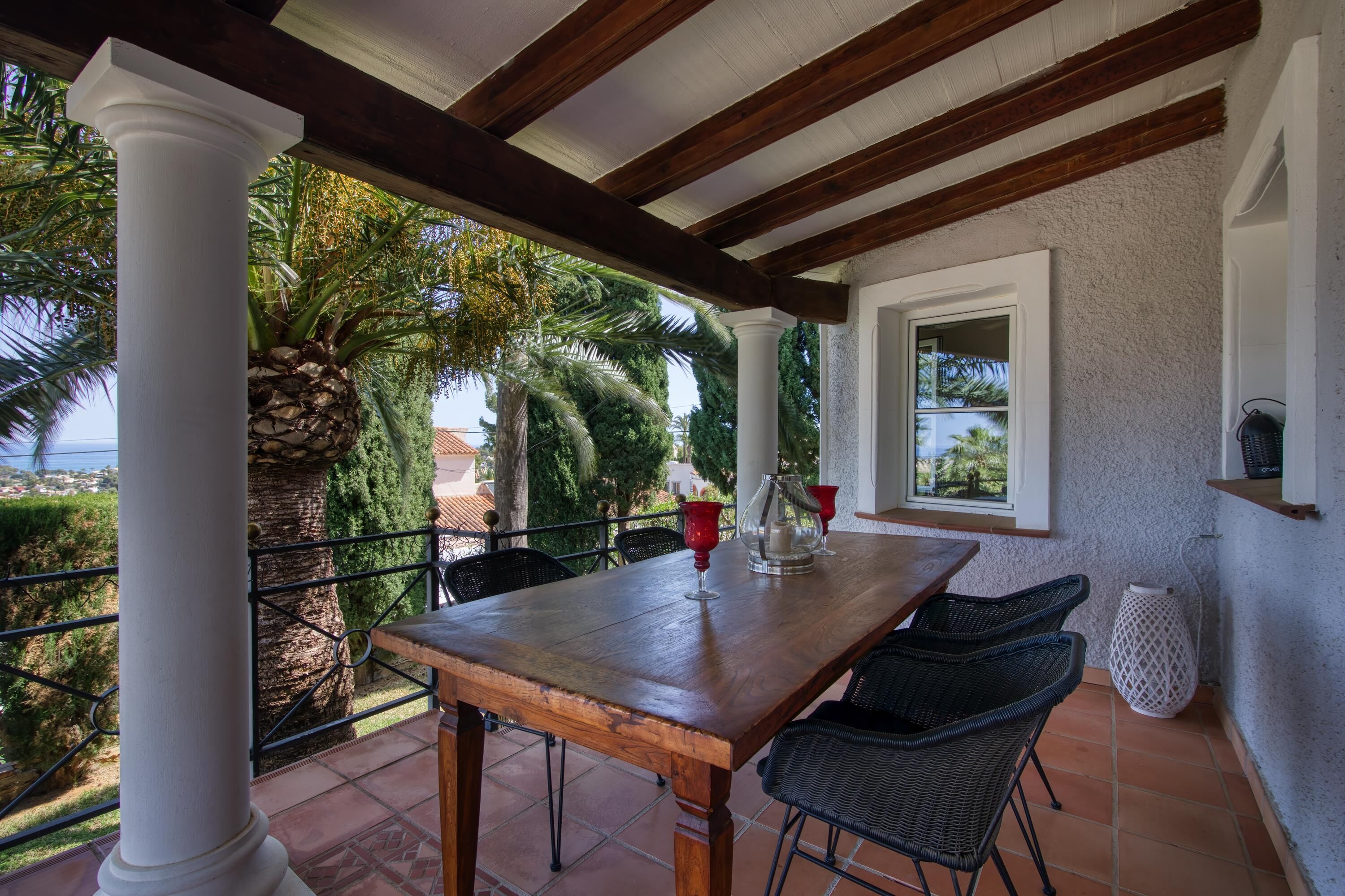 A covered patio area with a wooden table and chairs, a white column, and a view of palm trees and a building in the background.
