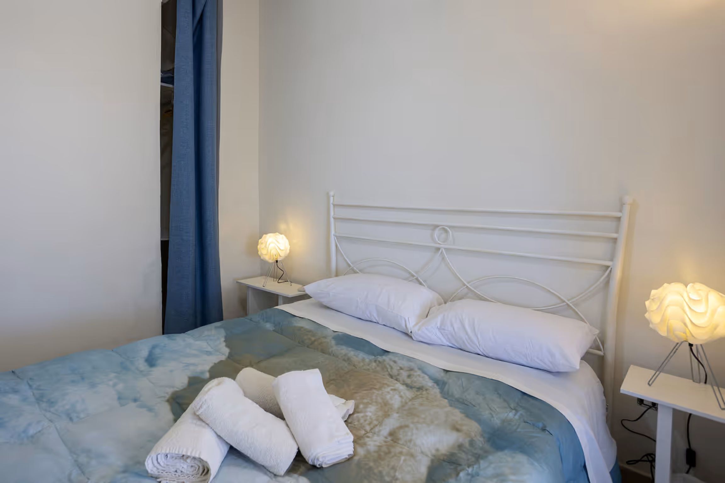 A neatly made bed with white linens and a blue and white patterned duvet, two bedside lamps, and a white nightstand with a decorative rose-shaped light.