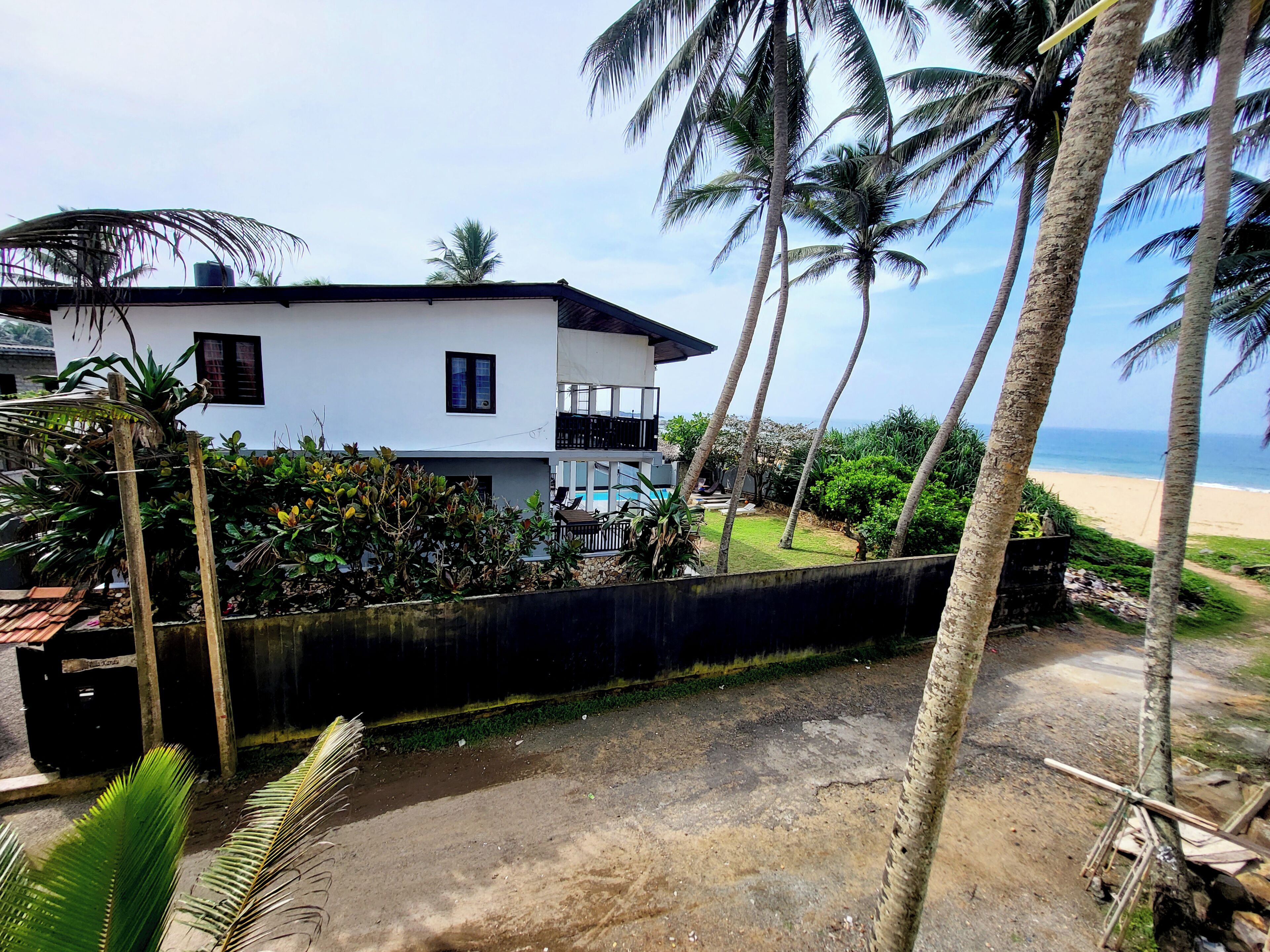 A white two-story house with a balcony, surrounded by palm trees and a view of the beach.