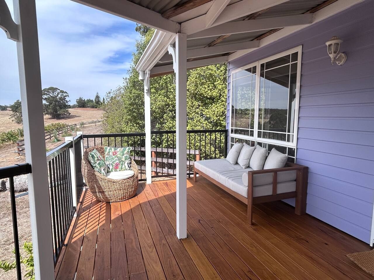 A wooden deck with a wicker chair and a sofa, a white railing, and a view of trees and hills.