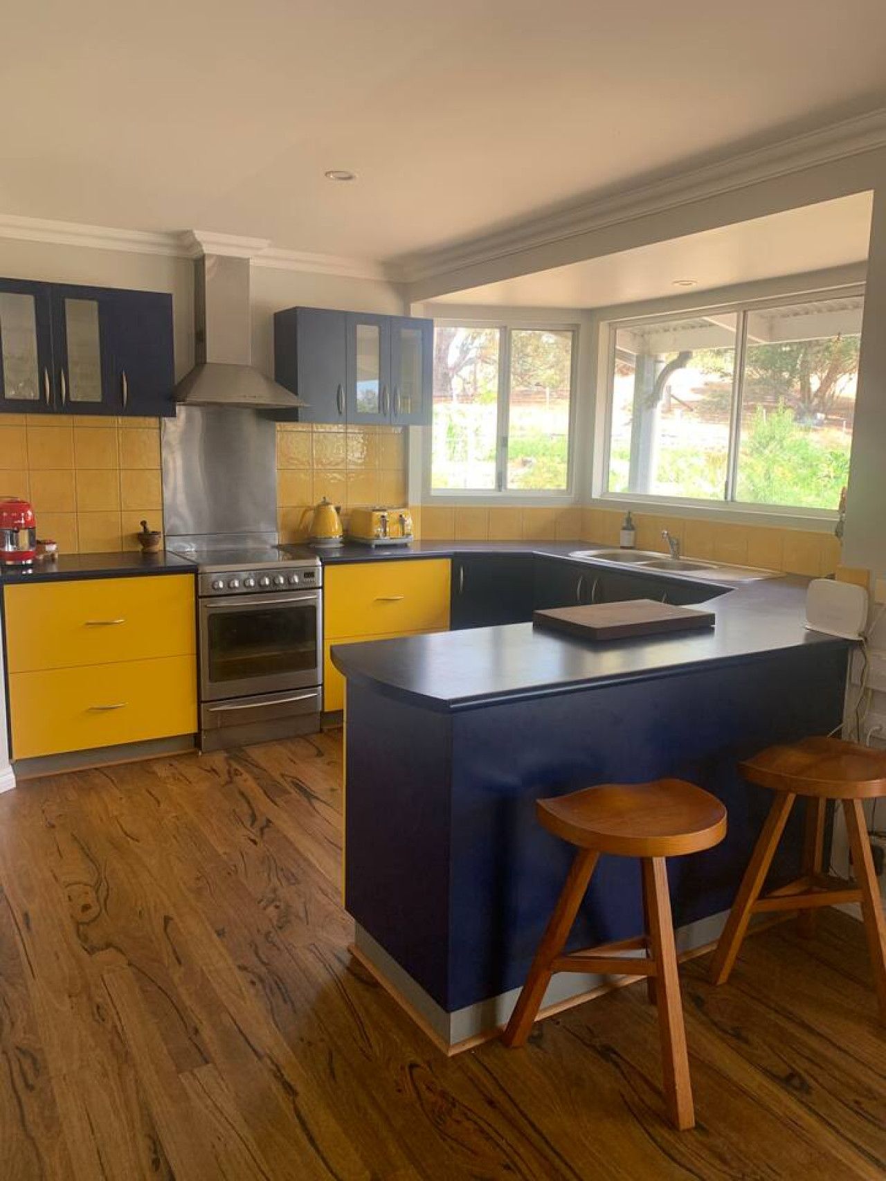 A modern kitchen with a central island, yellow and blue cabinetry, and a large window providing a view of the outdoors.