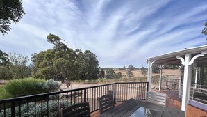 Une terrasse en bois avec une table en verre et des bancs, offrant une vue sur un paysage rural composé d’arbres et de champs ouverts.
