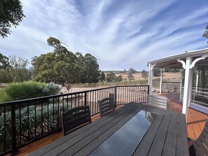 Una terraza de madera con una mesa de vidrio y bancos, con vistas a un paisaje rural con árboles y campos abiertos.