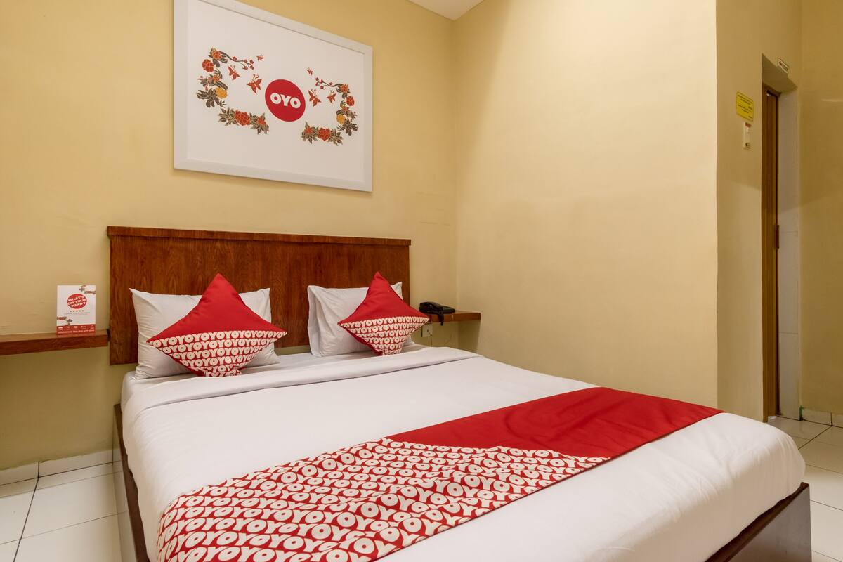 Ein Hotelzimmer mit einem Bett, einem hölzernen Kopfteil, roten und weißen Kissen, einer roten und weißen Tagesdecke und einem gerahmten Bild an der Wand.