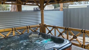 A hot tub under a bamboo roof structure with a wooden bench and a metal fence in the background.