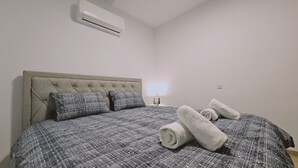 A bedroom with a tufted headboard, a bed with a patterned duvet, two pillows, a bedside lamp, and rolled towels on the bed. - STAY Cosmo City Suite 303 (Nicosia)