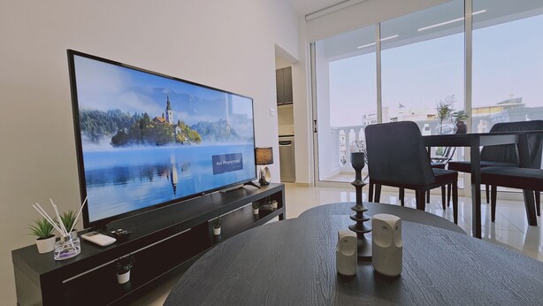 A modern living room with a large flat-screen TV on a black stand, a dining area with a round table and chairs, and a view of the city through large windows. - STAY Cosmo City Suite 303 (Nicosia)