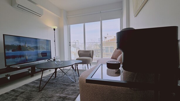 A modern living room with a large flat-screen TV, a glass coffee table, a comfortable sofa, and a chair. - STAY Cosmo City Suite 102 (Nicosia)