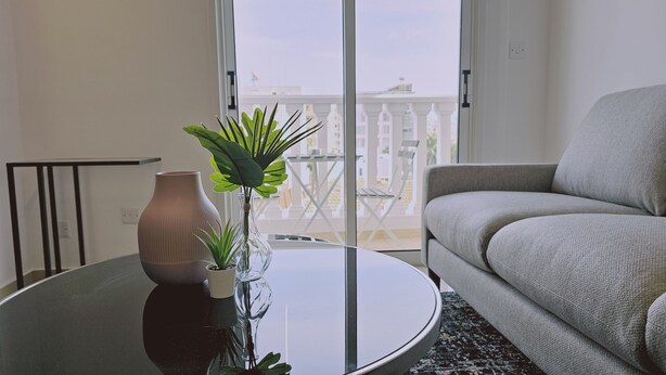 A modern living room with a round glass coffee table, a grey sofa, and a potted plant. - STAY Cosmo City Suite 302 (Nicosia)