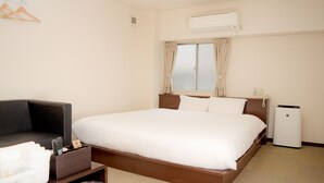 Een hotelkamer met een bed, een klein tafeltje met een prullenbak, een stoel en een airconditioner.