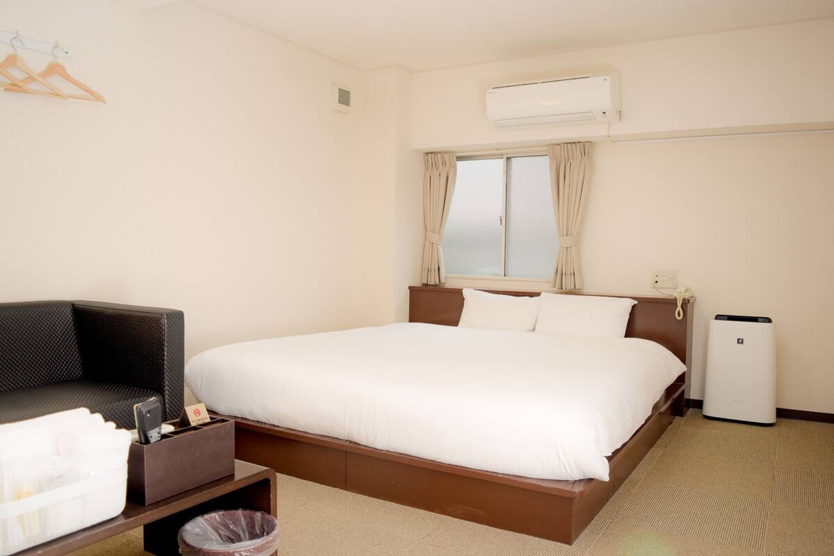 Habitación de hotel con cama, una mesita de noche con un cubo de basura, una silla y un equipo de aire acondicionado.
